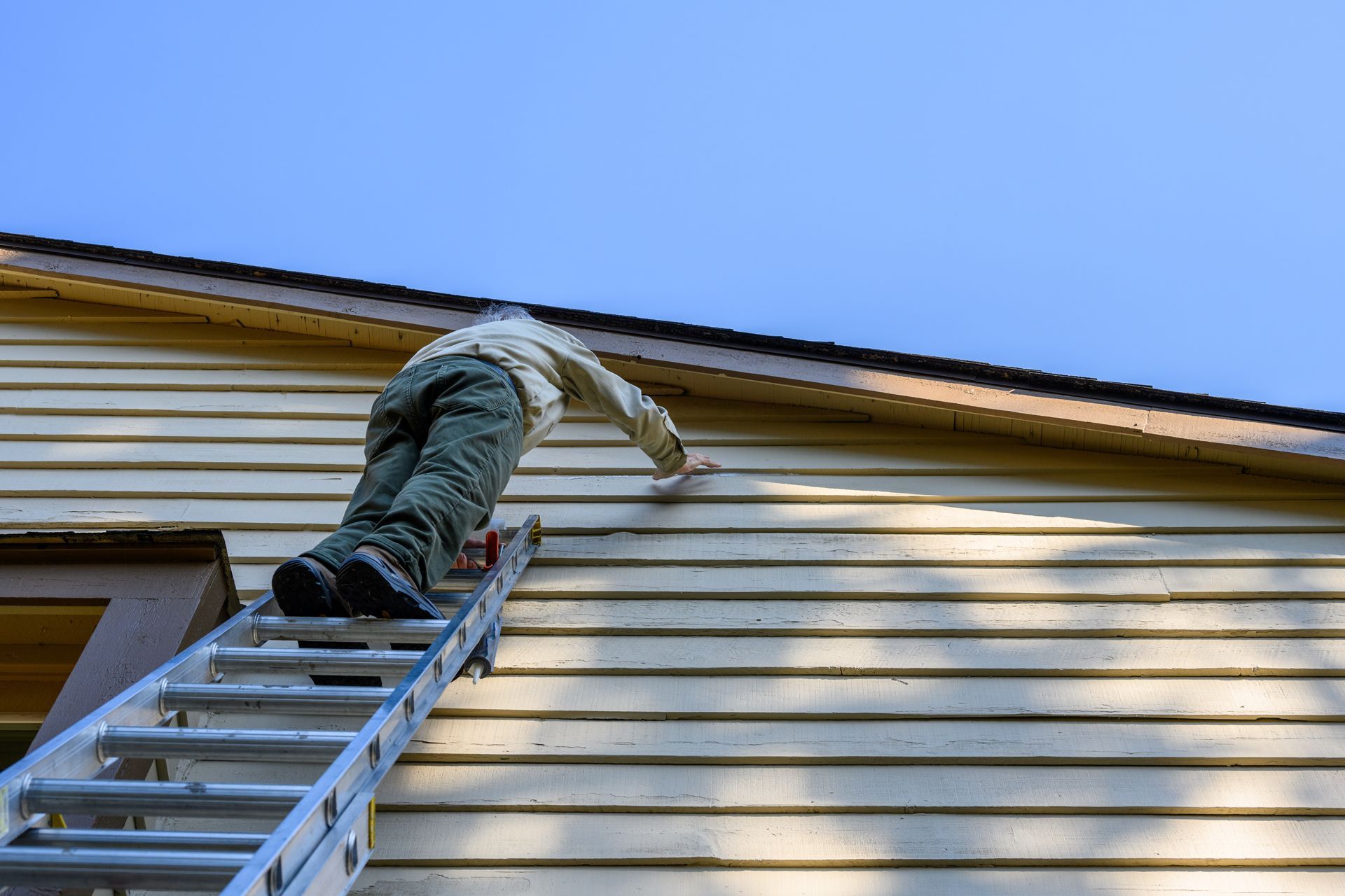 Person painting the side of a house from a ladder, under a blue sky.