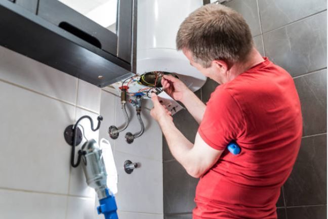 Un homme en chemise rouge répare un chauffe-eau dans une salle de bain.