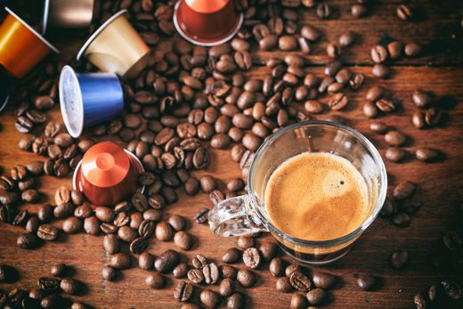 Chicchi di caffè sparsi intorno a tazzine da espresso e una tazza di caffè in vetro su un tavolo di legno.