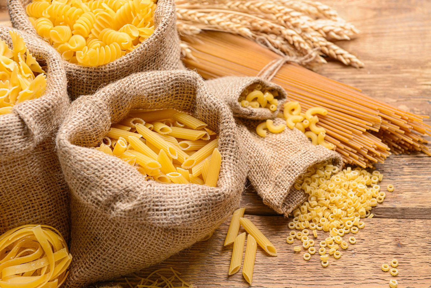 Assortimento di pasta in sacchi di iuta e sparsa su un tavolo di legno con spighe di grano.