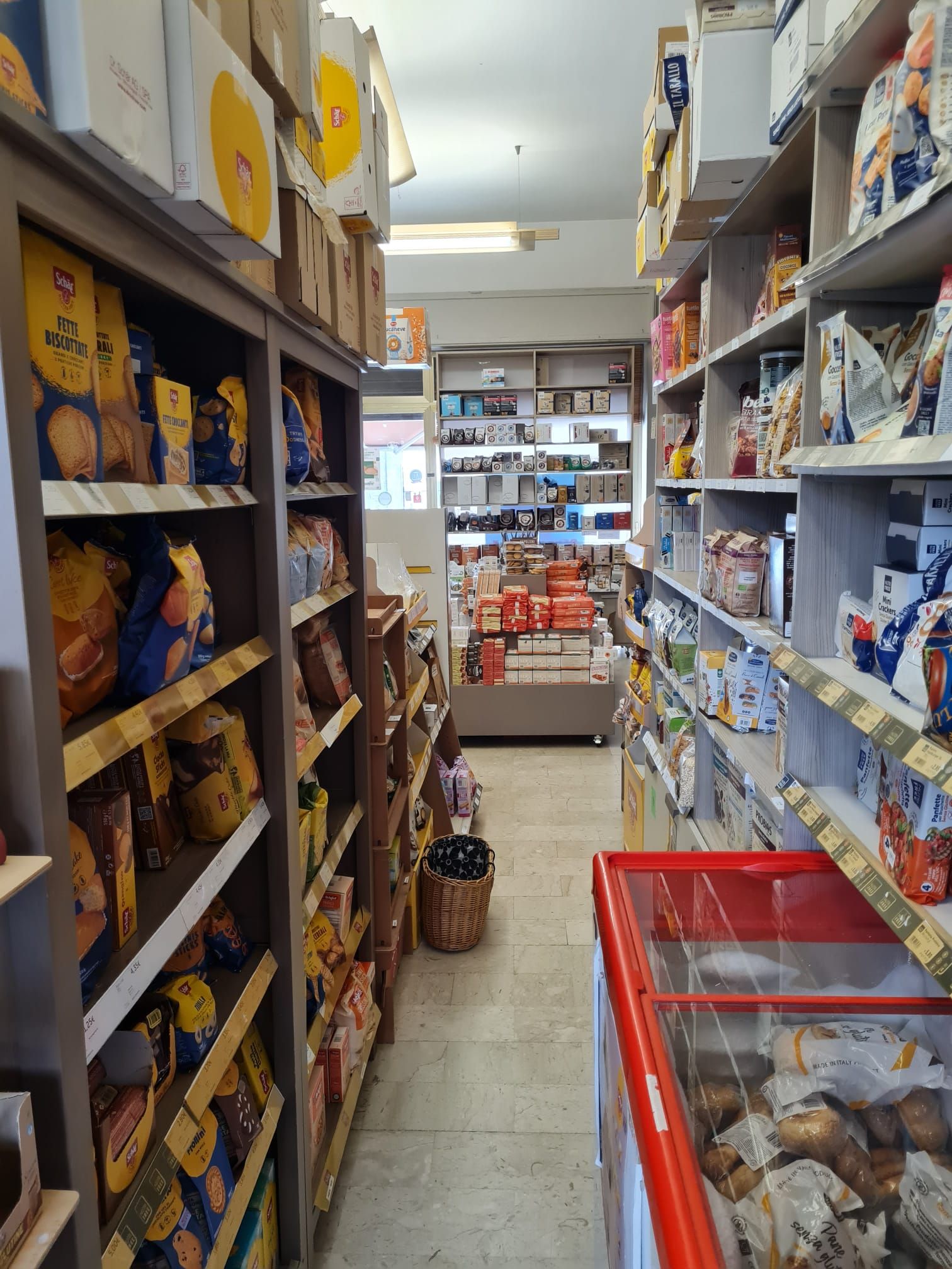 Una corsia di un supermercato con scaffali pieni di pane e snack, e un carrello della spesa rosso in primo piano.