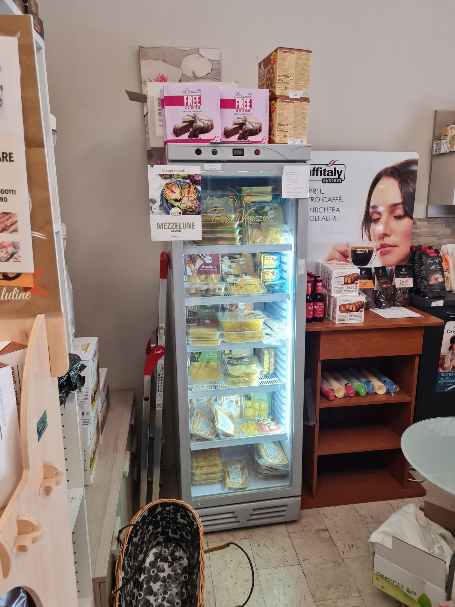 Un piccolo frigorifero da negozio, rifornito di dolci e bevande, si trova accanto a scaffali e scatole in un interno angusto.