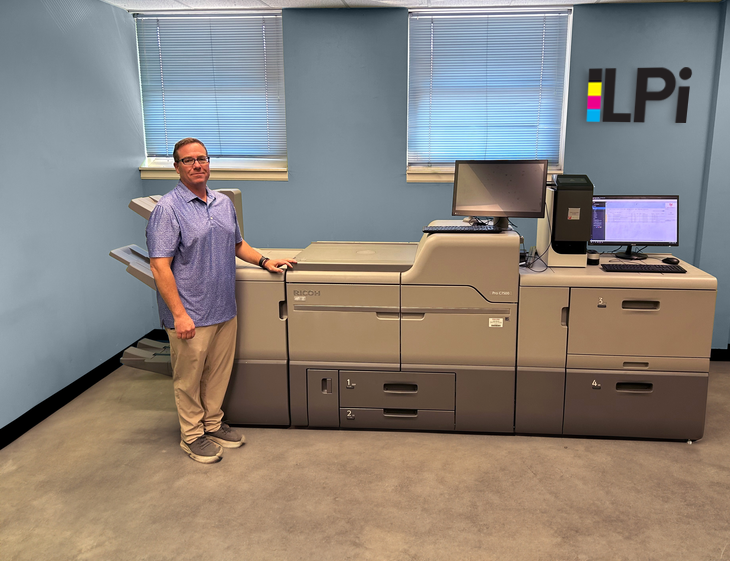 Man stands beside large printing machine in an office. The background is blue.