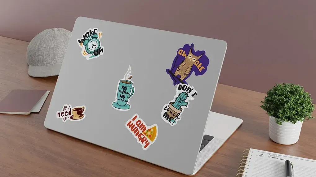 Laptop with stickers on a wooden desk next to a potted plant, notebook, and a hat.