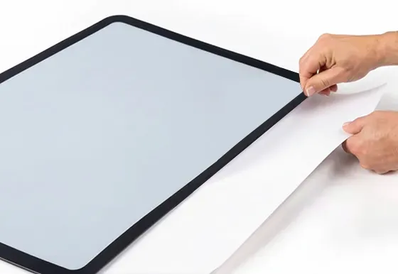Hands peeling a white protective film from a rectangular screen with a black frame.