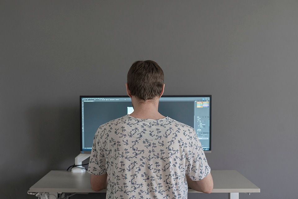 Person facing a computer monitor, working at a desk, back to the camera, against a gray wall.
