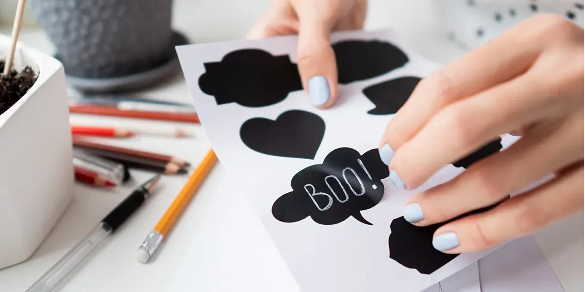 Hands arranging black cloud-shaped paper cutouts and a “BOO!” label on a white desk