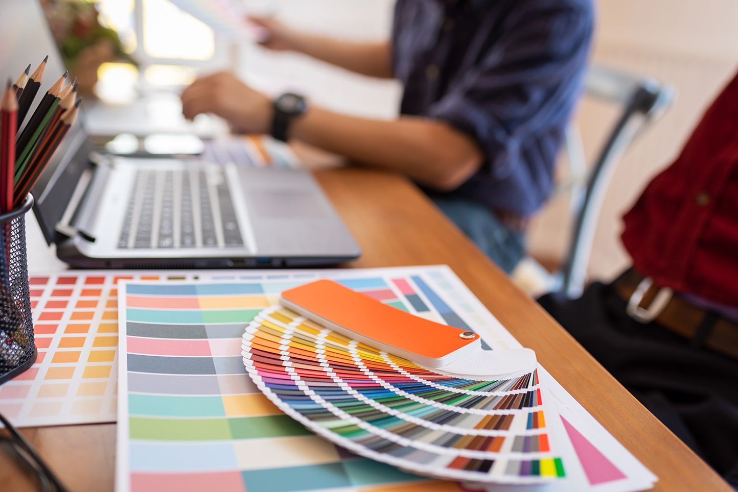 Desk with a laptop, color swatches, and a person holding papers, focused on design work.