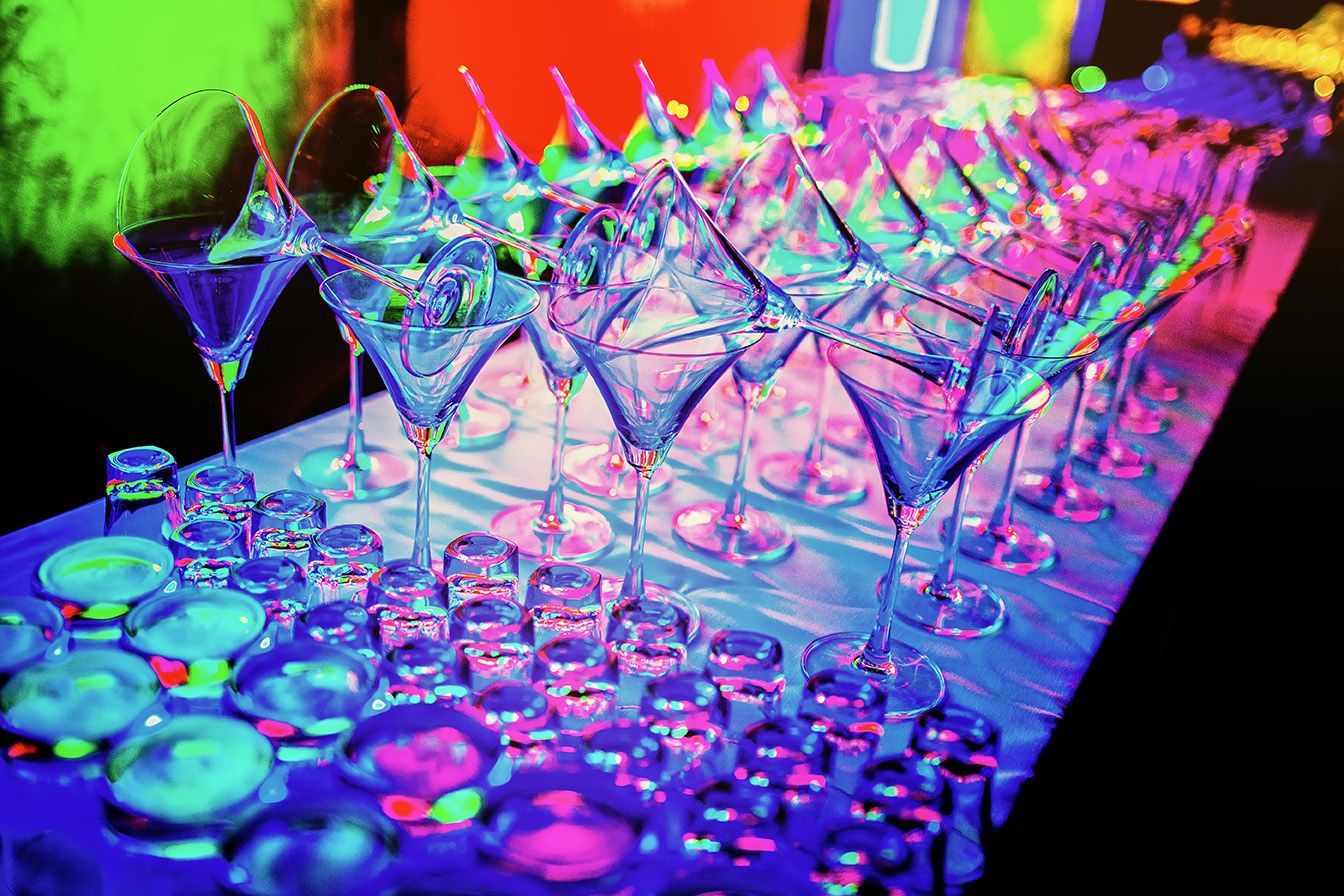 Glowing martini glasses and shot glasses on a bar under neon lights.