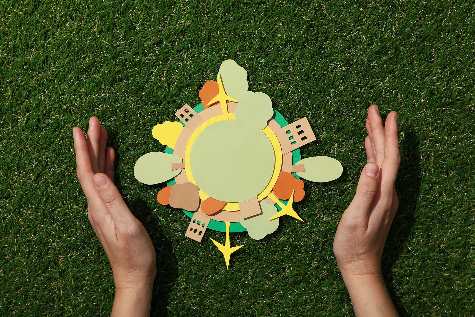 Hands cupping a paper cutout globe with buildings, trees, and airplanes on a grassy background.