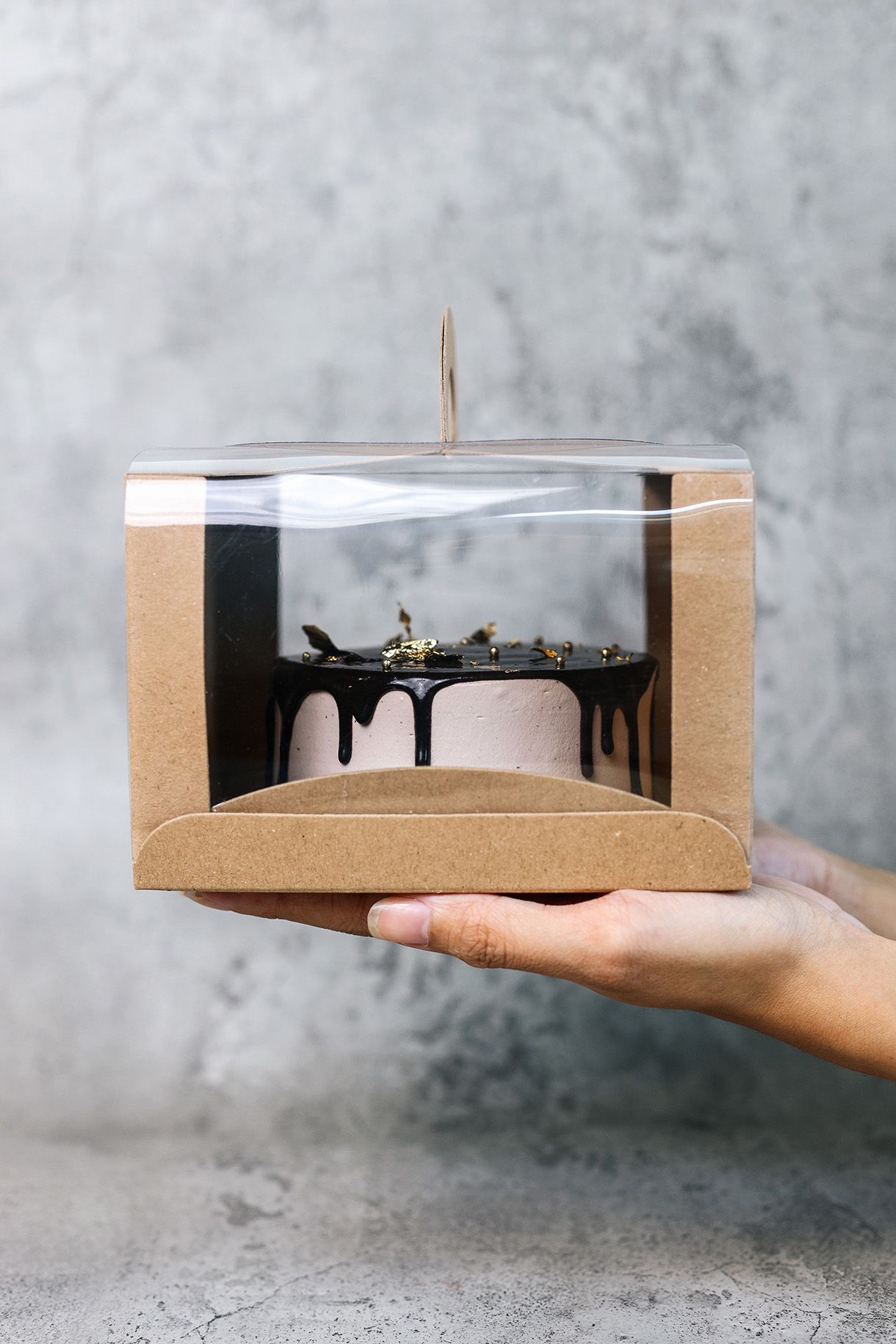 Person holding a chocolate cake in a clear-topped brown box against a gray backdrop. The cake has chocolate drips.