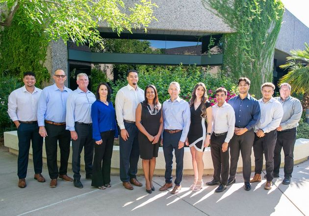 A group of thirteen people standing in a line outdoors in front of a building with ivy-covered walls and bright sunlight.