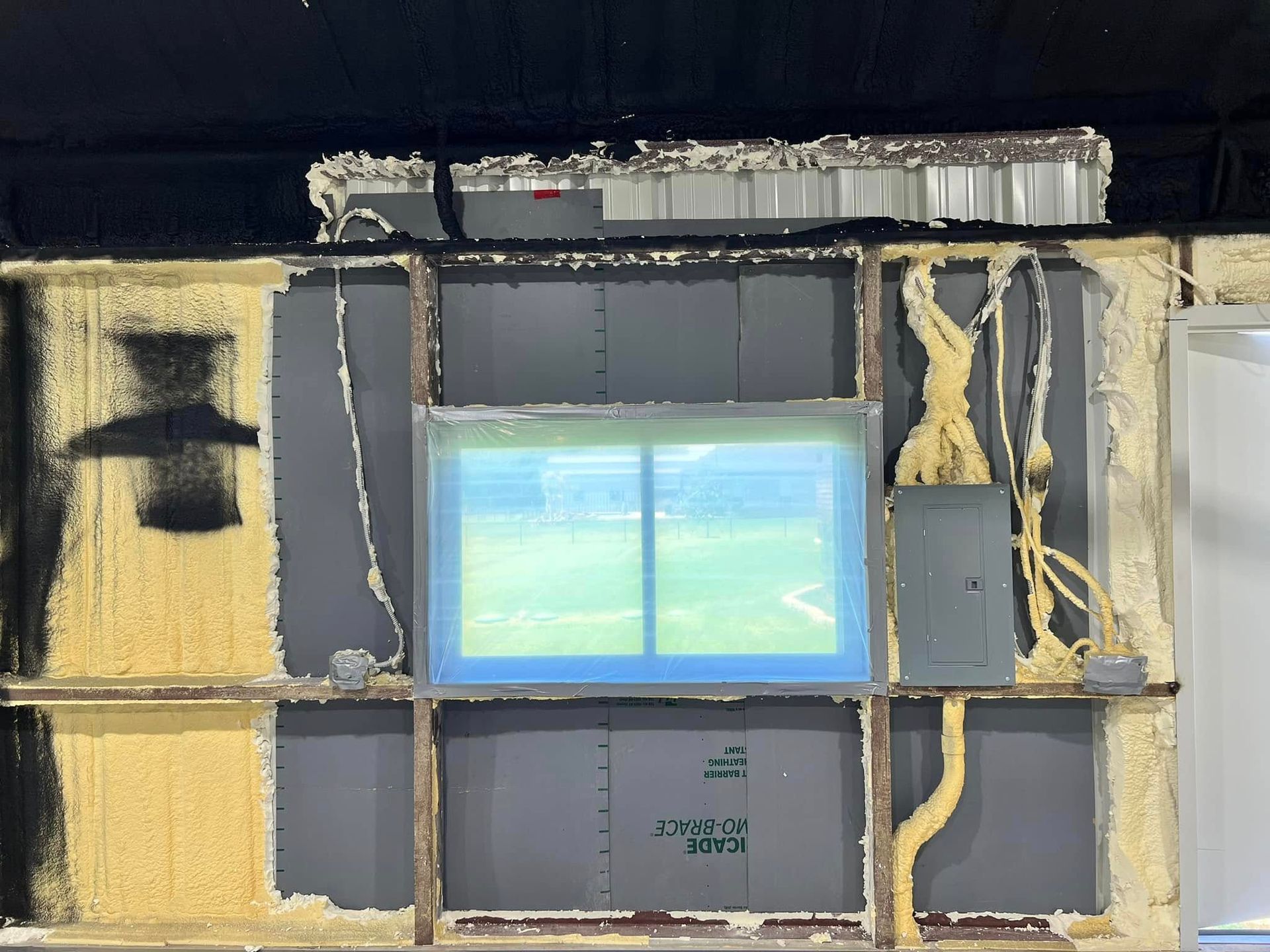 Wall construction with a window, partially filled with yellow insulation and gray panels, with a view of a green field.