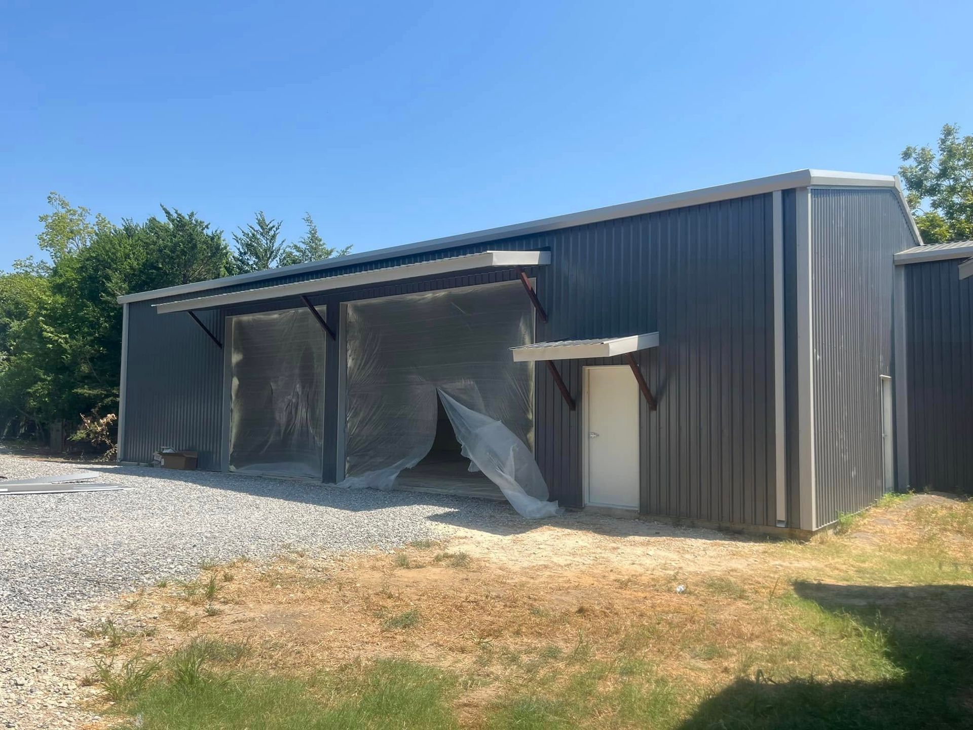 Gray metal shed with a gravel drive, white door, and clear plastic sheeting.