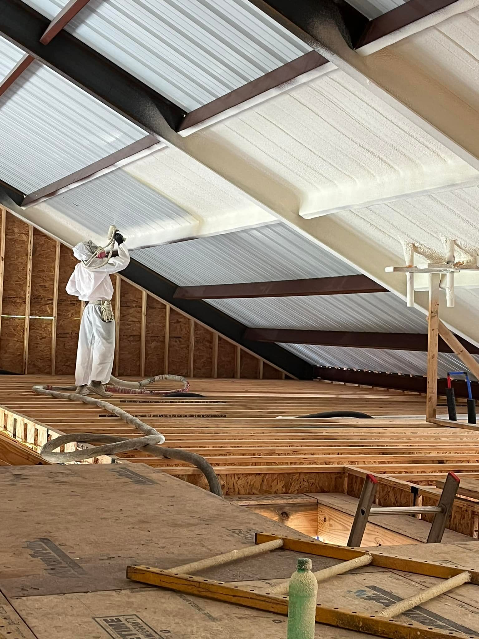 Worker spraying foam insulation onto a roof. Construction setting with exposed wood beams and metal roofing.