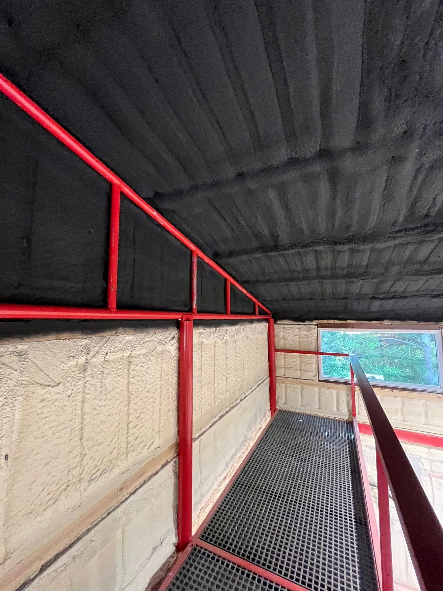 Red railing on a metal grated walkway, adjacent to a window, with a black foam-coated ceiling.
