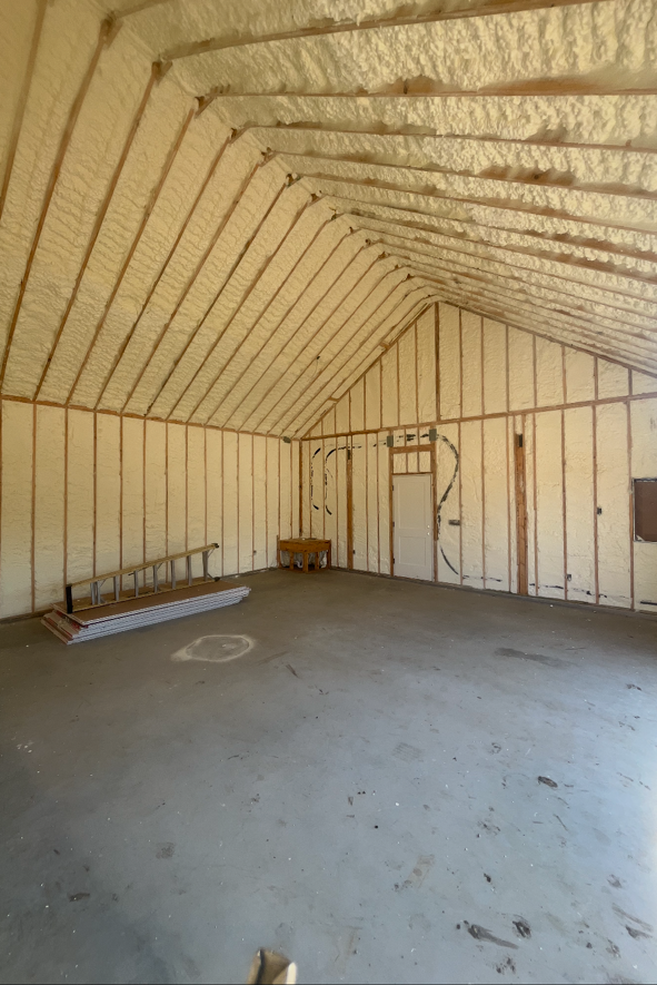 Interior view of a building with spray foam insulation on walls and ceiling, empty concrete floor, and wooden beams.