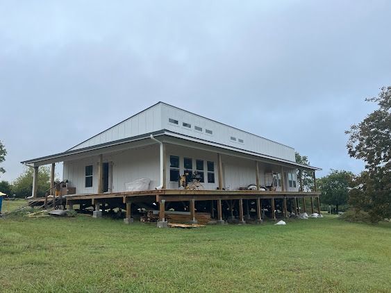 White house with wrap-around porch under construction. Green grass, cloudy sky, support beams visible.