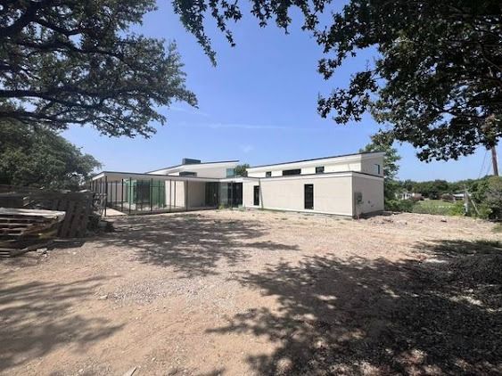 Modern beige house with flat roof, glass walls, and a gravel yard. Trees frame the shot under a blue sky.