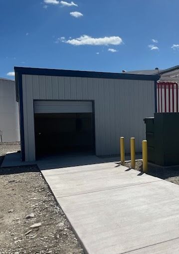Exterior of a light gray metal shed with open garage door. Concrete path leads to entrance. Blue sky.