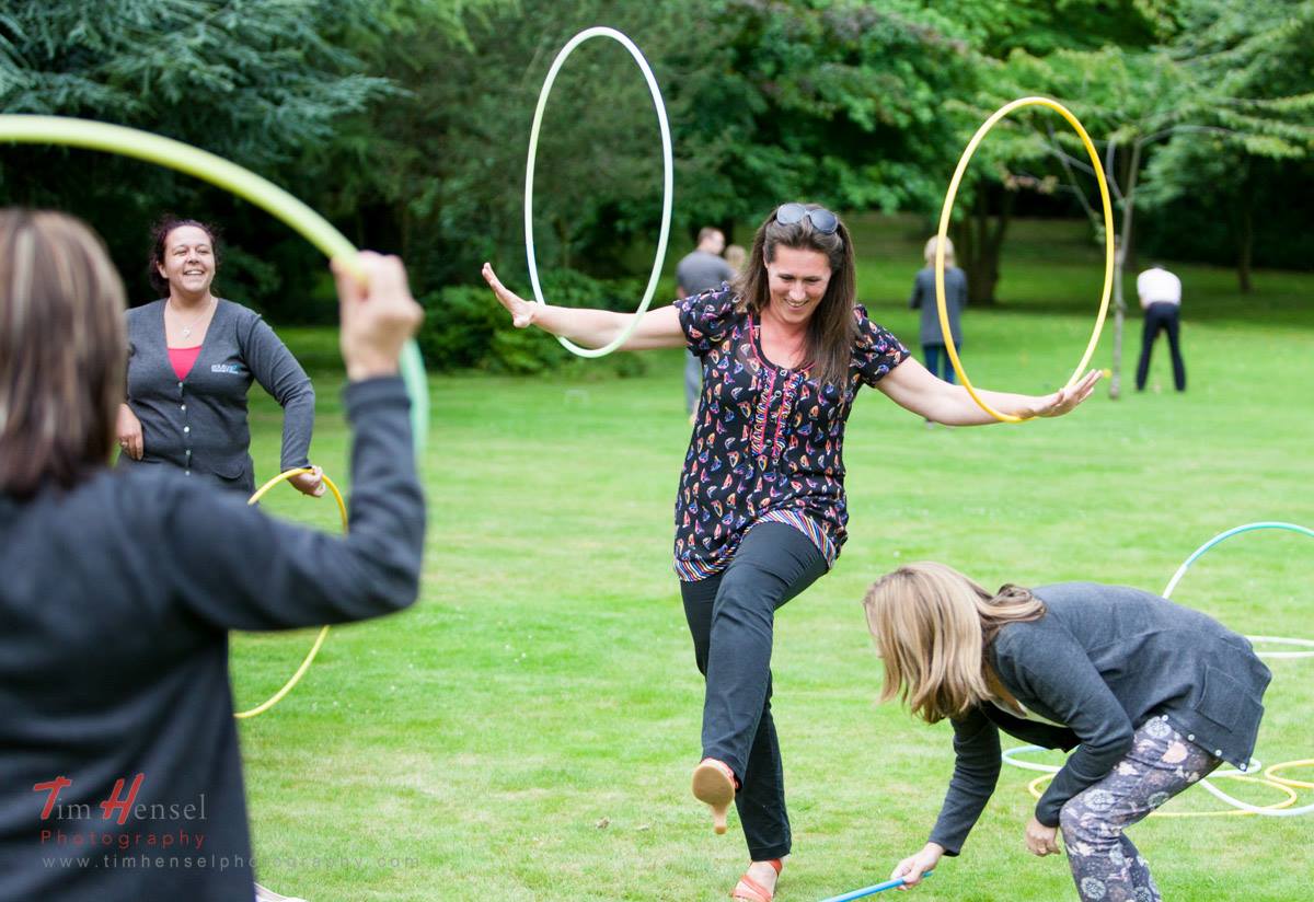 People playing with hula hoops outdoors on a grassy lawn. One woman is balancing a hoop while smiling. Giggles & Games