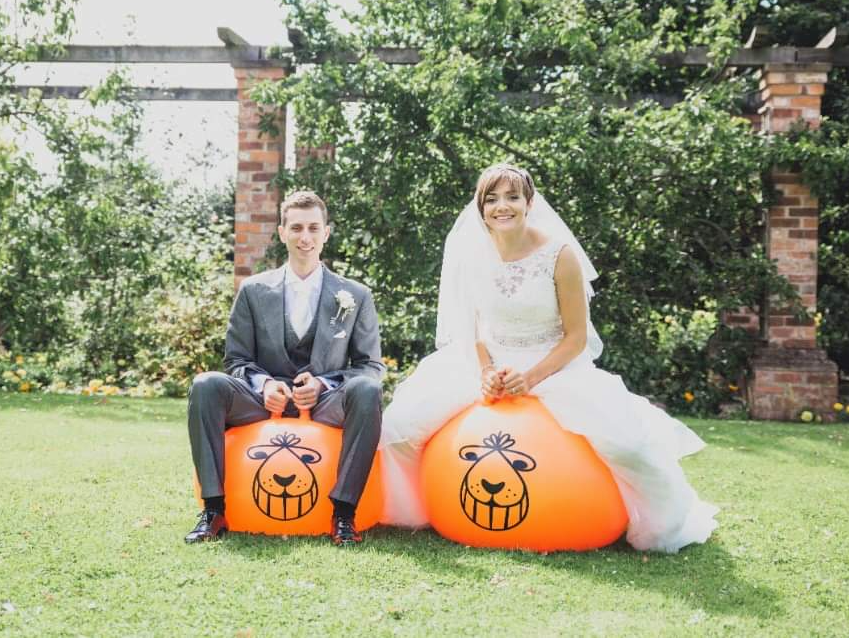 Bride and groom on orange space hoppers with grinning face design, in a garden. Giggles & Games
