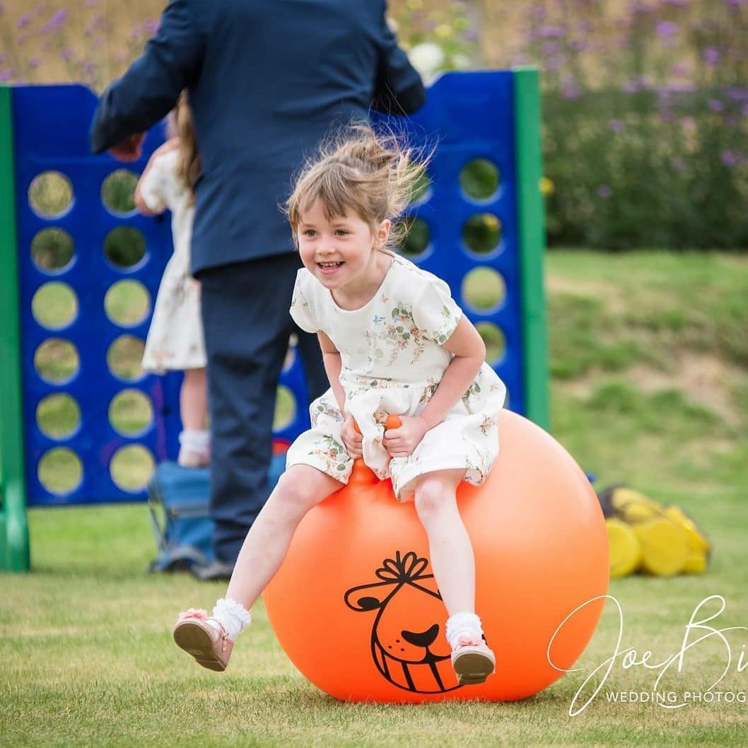 Girl bounces on an orange hopper ball in a grassy area, smiling. A Connect Four game is behind her. Wedding Games