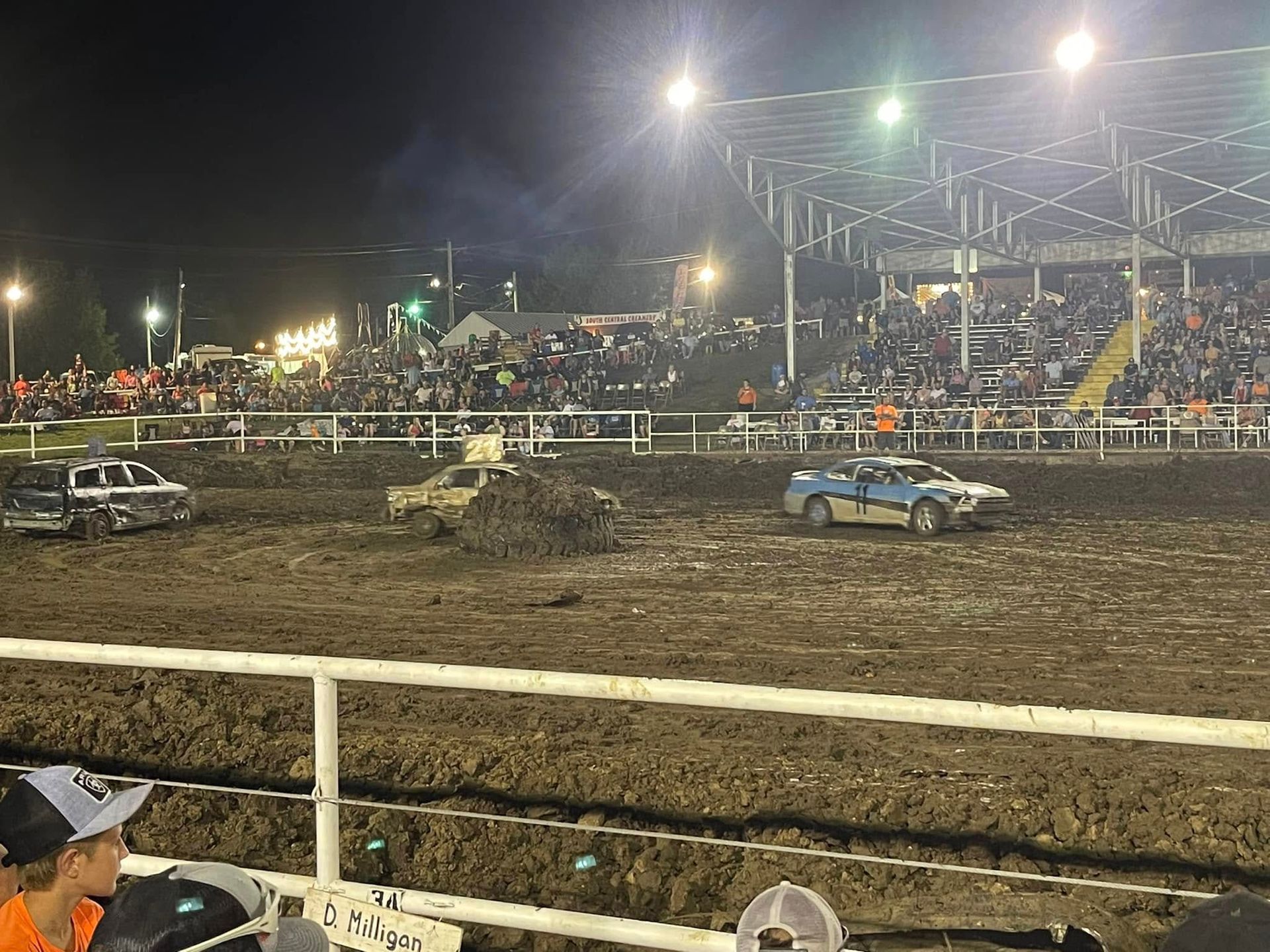 Mud-filled arena with two derby cars racing at night under bright lights, crowd in bleachers.
