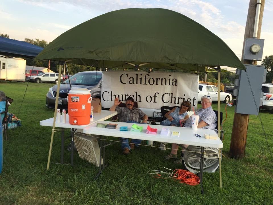 California Church of Christ outreach booth under a green canopy with volunteers seated at a table.