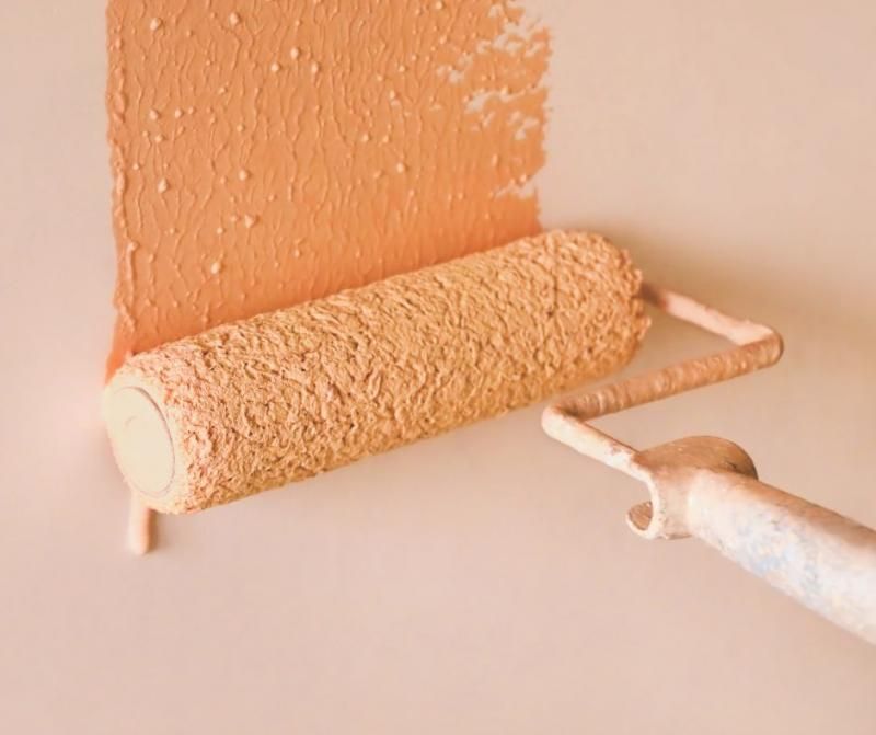 A paint roller applies a thick layer of peachy-orange paint onto a solid, lighter-toned wall surface.
