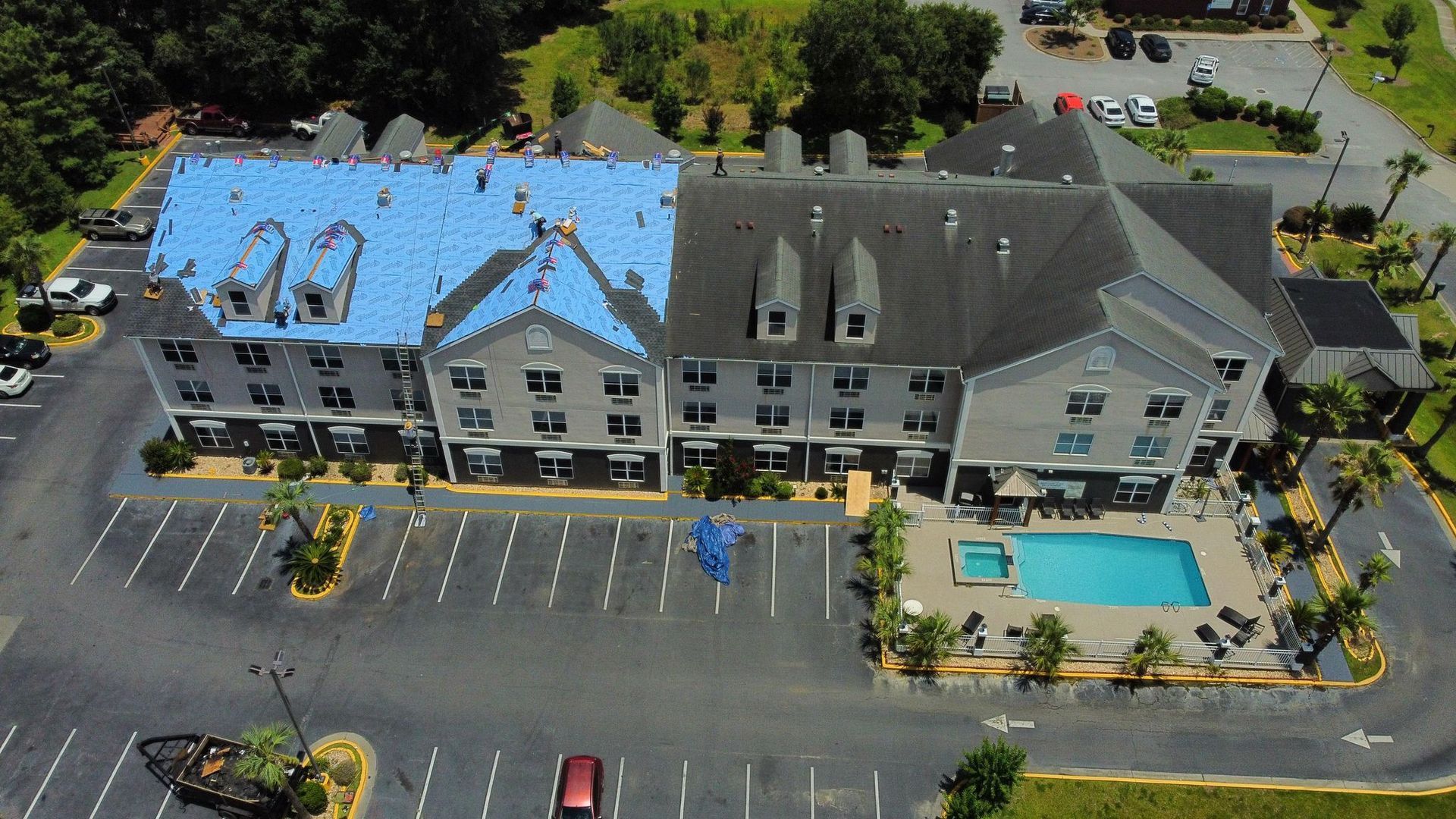Aerial view of a hotel with blue roof shingles being installed, pool, and parking lot.