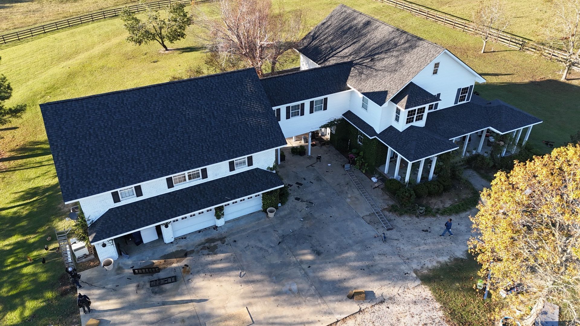Large white house with black roof, a large concrete driveway, and surrounding green fields.