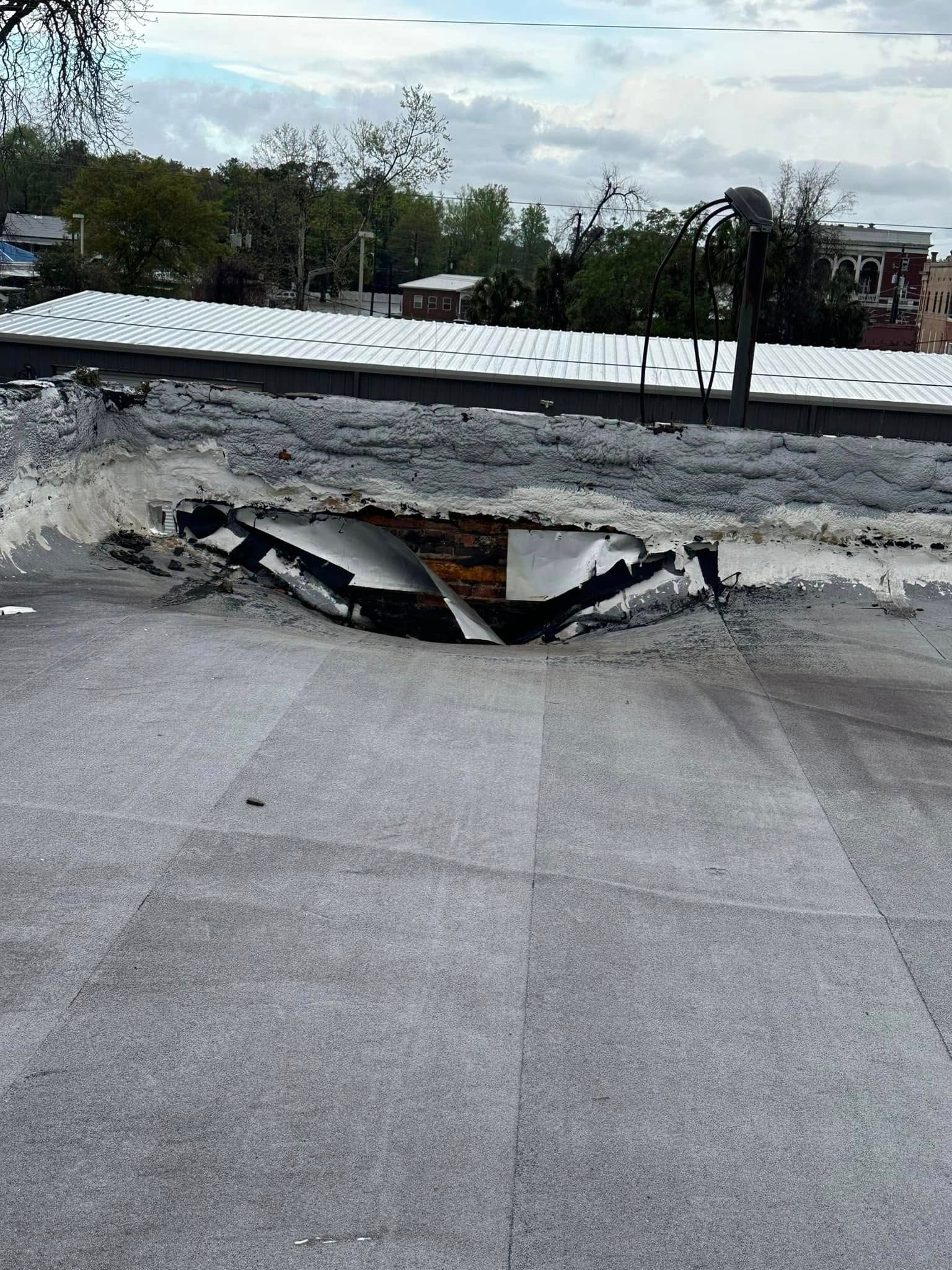 Damaged flat roof with a large hole; trees and buildings in background.
