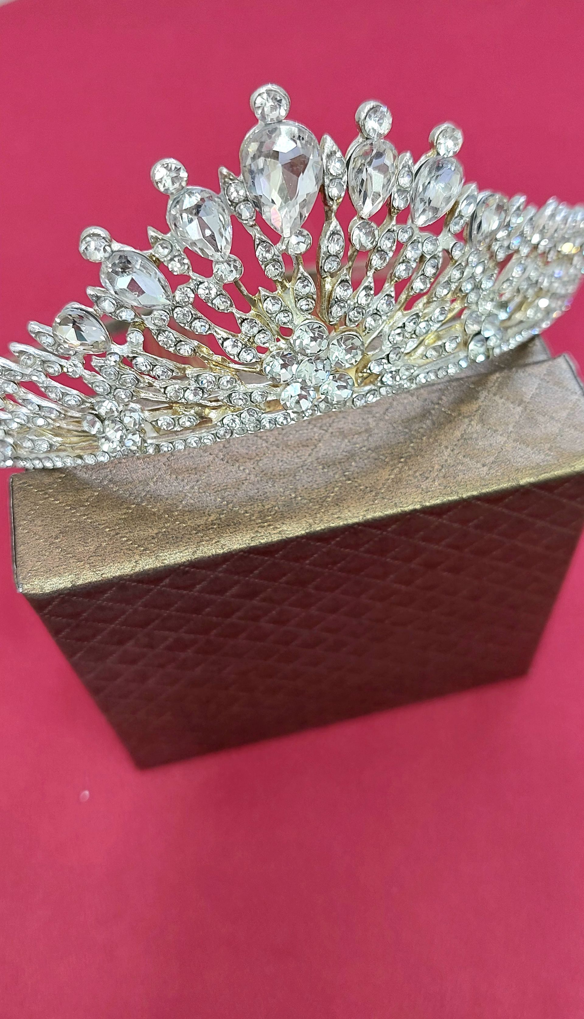 Tiara in argento con gioielli trasparenti su una scatola con texture dorata su sfondo rosso.
