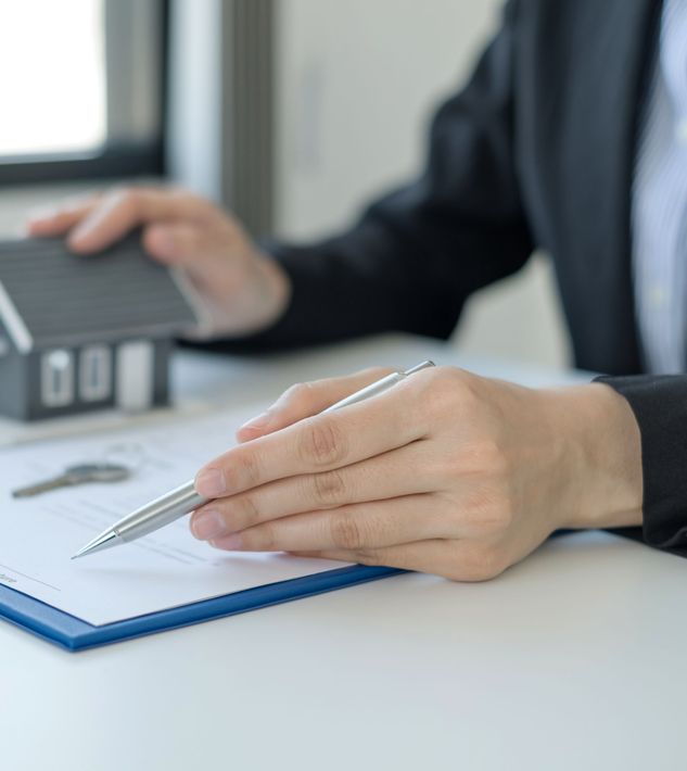 A person is holding a pen over a clipboard with a model house in the background.