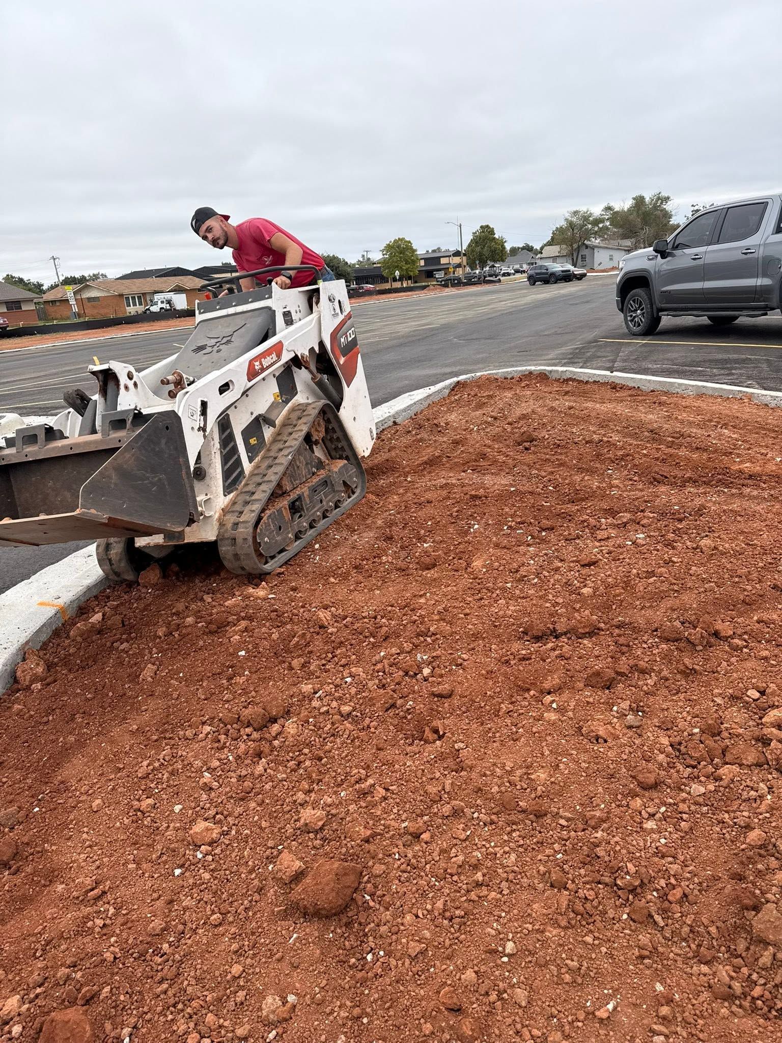 Worker operating a skid steer to spread mulch along a roadside curb under cloudy skies