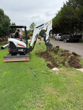 Mini excavator digging a trench in a grassy yard beside a road on a cloudy day