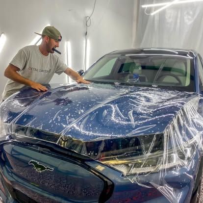 A man is covering the hood of a blue mustang with plastic wrap.