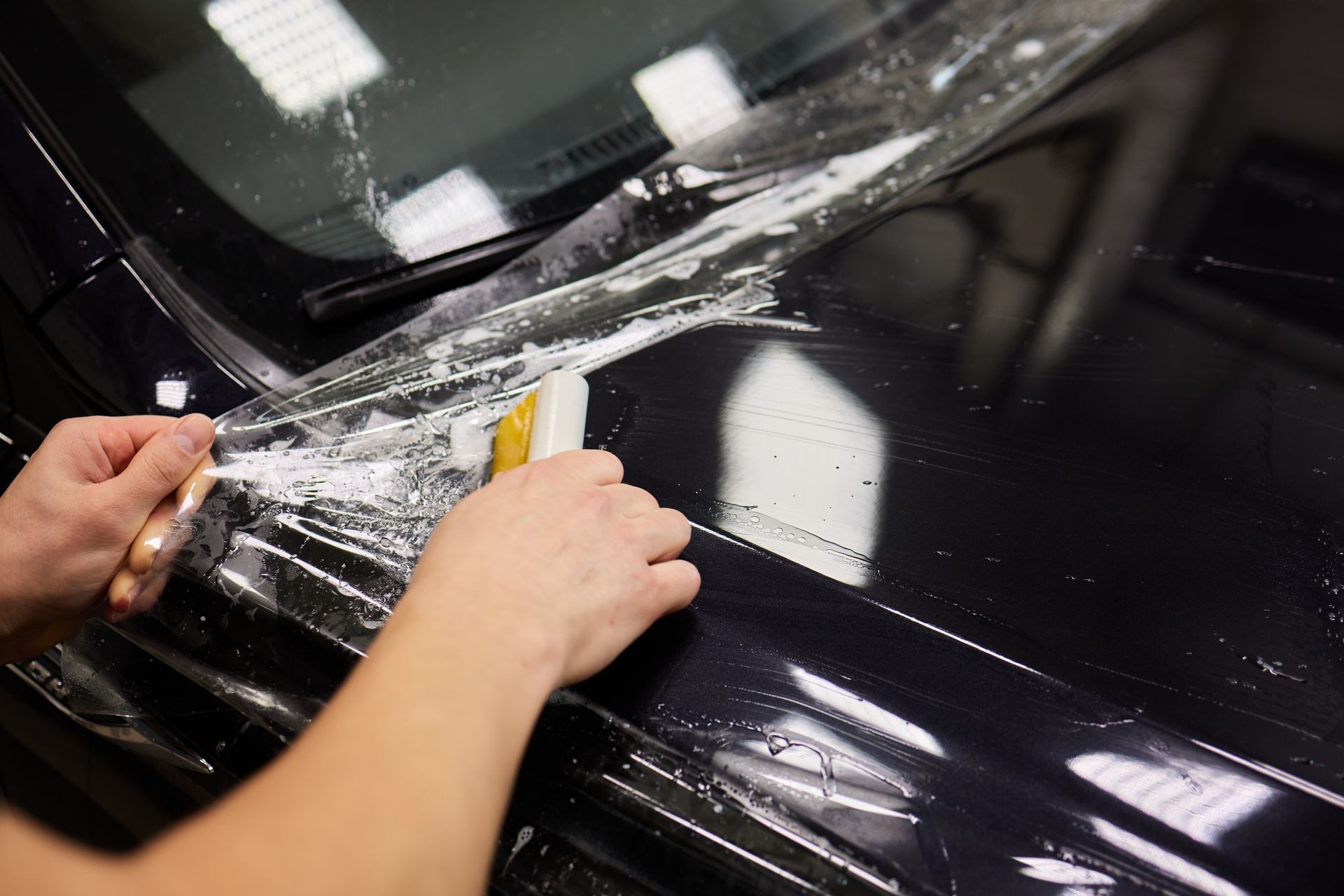 Hands applying clear protective film to a black car hood with a squeegee