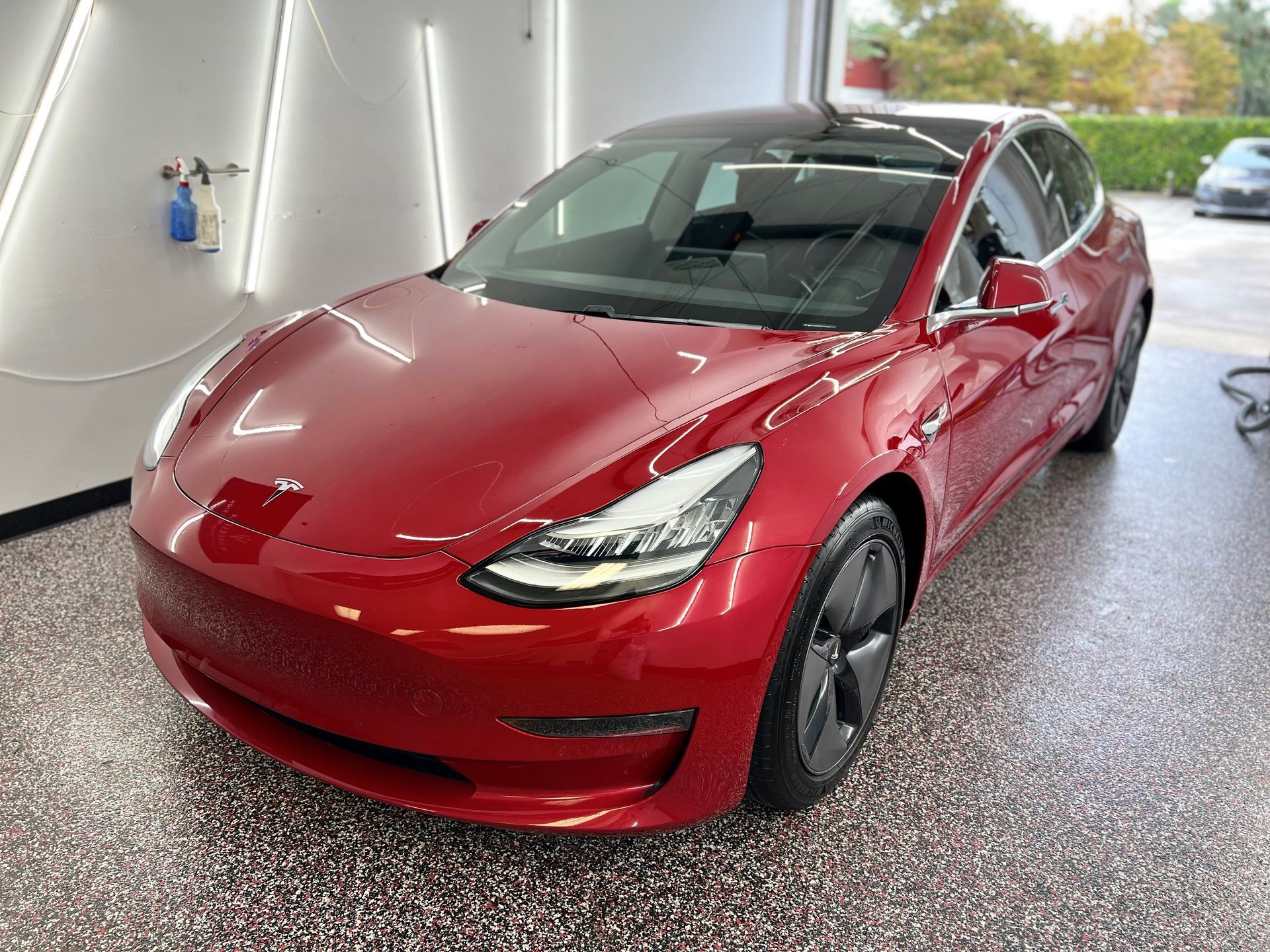 Red Tesla Model 3 parked in a garage, with black rims and roof, shiny paint.