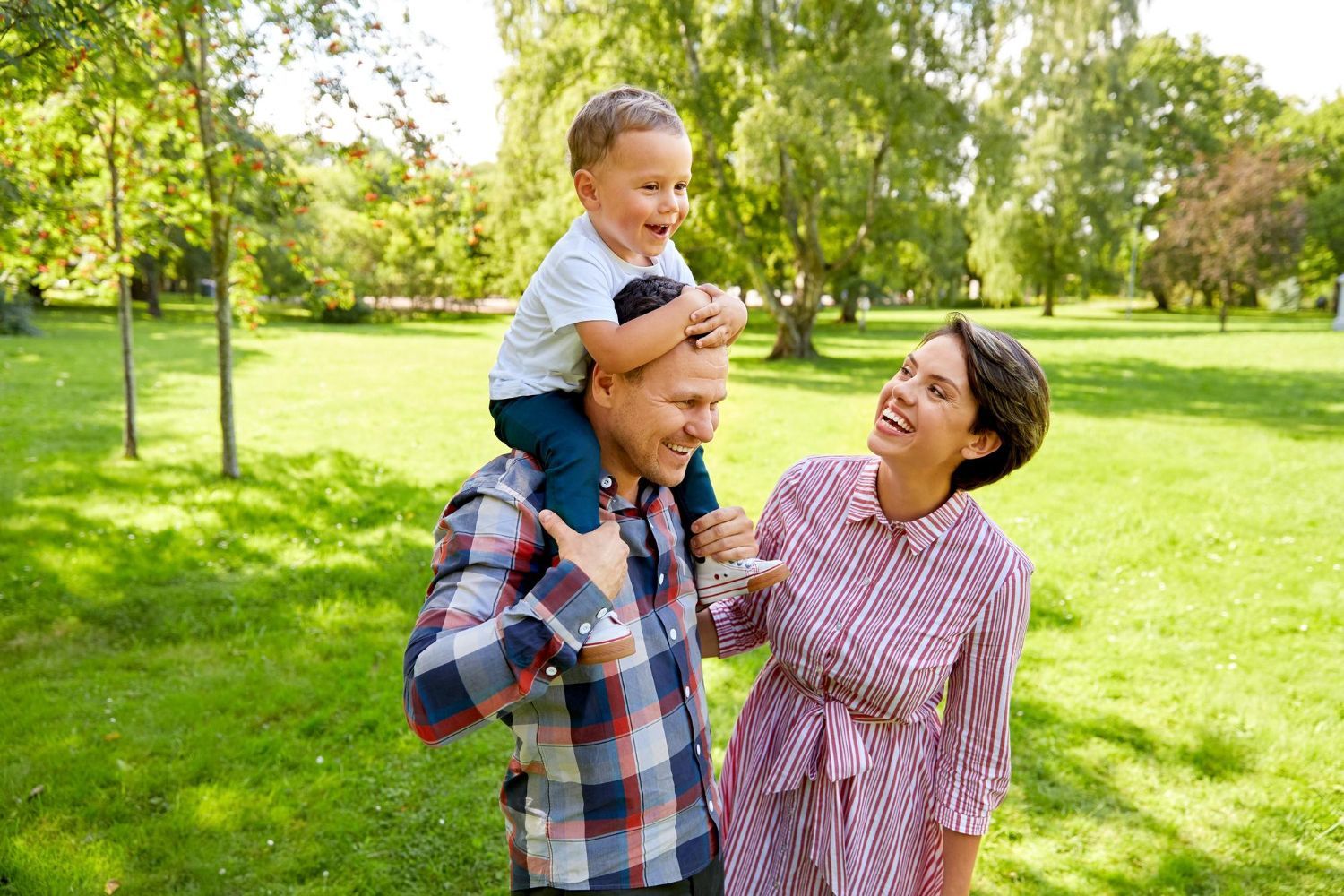 Adult carrying a smiling child on shoulders in a sunny green park, with a woman walking beside them