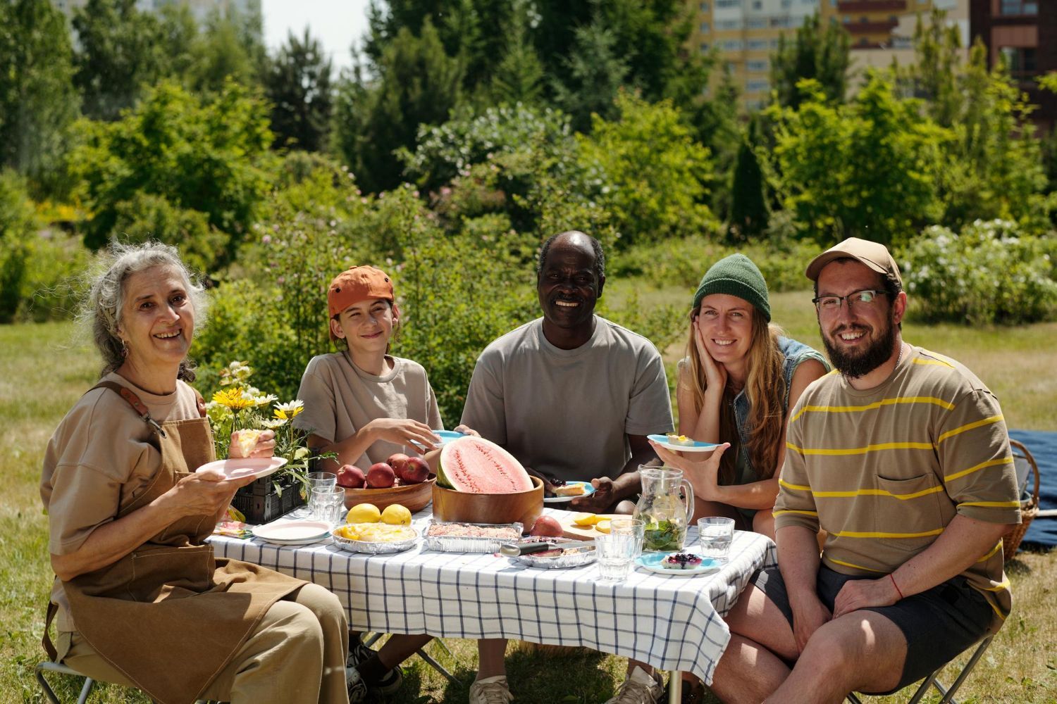 Five friends picnic at a table outdoors in a sunny green field, smiling and sharing food.