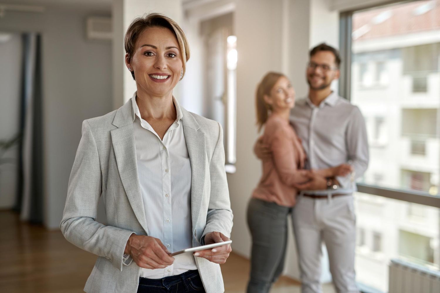 Smiling woman in a light blazer holding keys in a bright apartment with a couple in the background