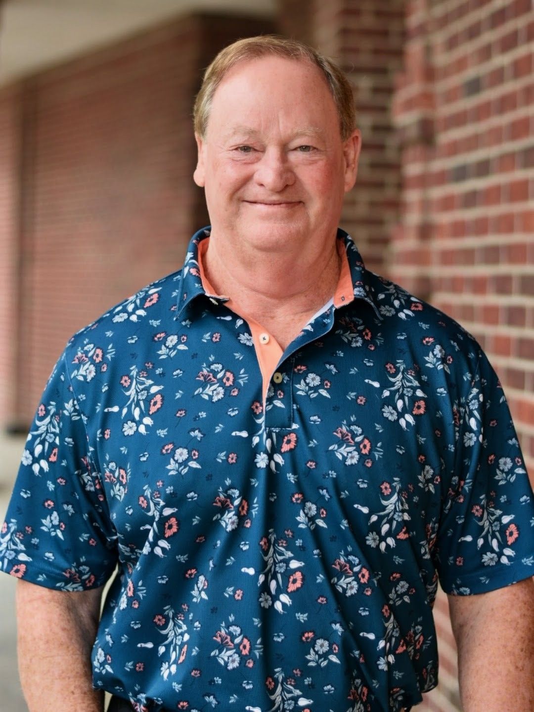 Man seated in an office chair, smiling, wearing a navy polo, with plants and a desk behind him.