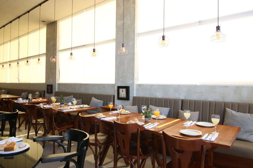 Sala de jantar vazia de um restaurante com mesas de madeira, cadeiras brancas e grandes janelas que deixam entrar muita luz.