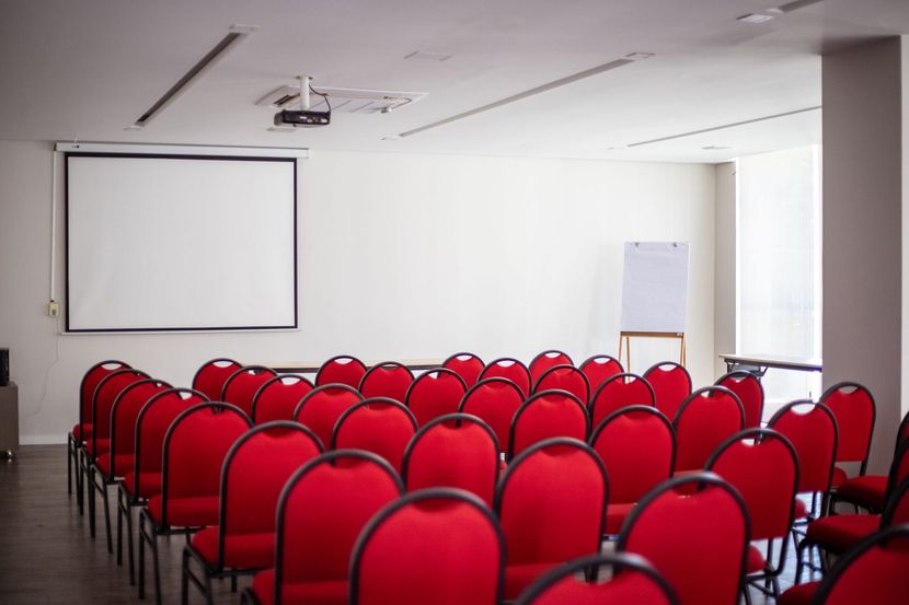 Sala de conferências vazia com fileiras de cadeiras vermelhas, uma tela de projeção e um flip chart.