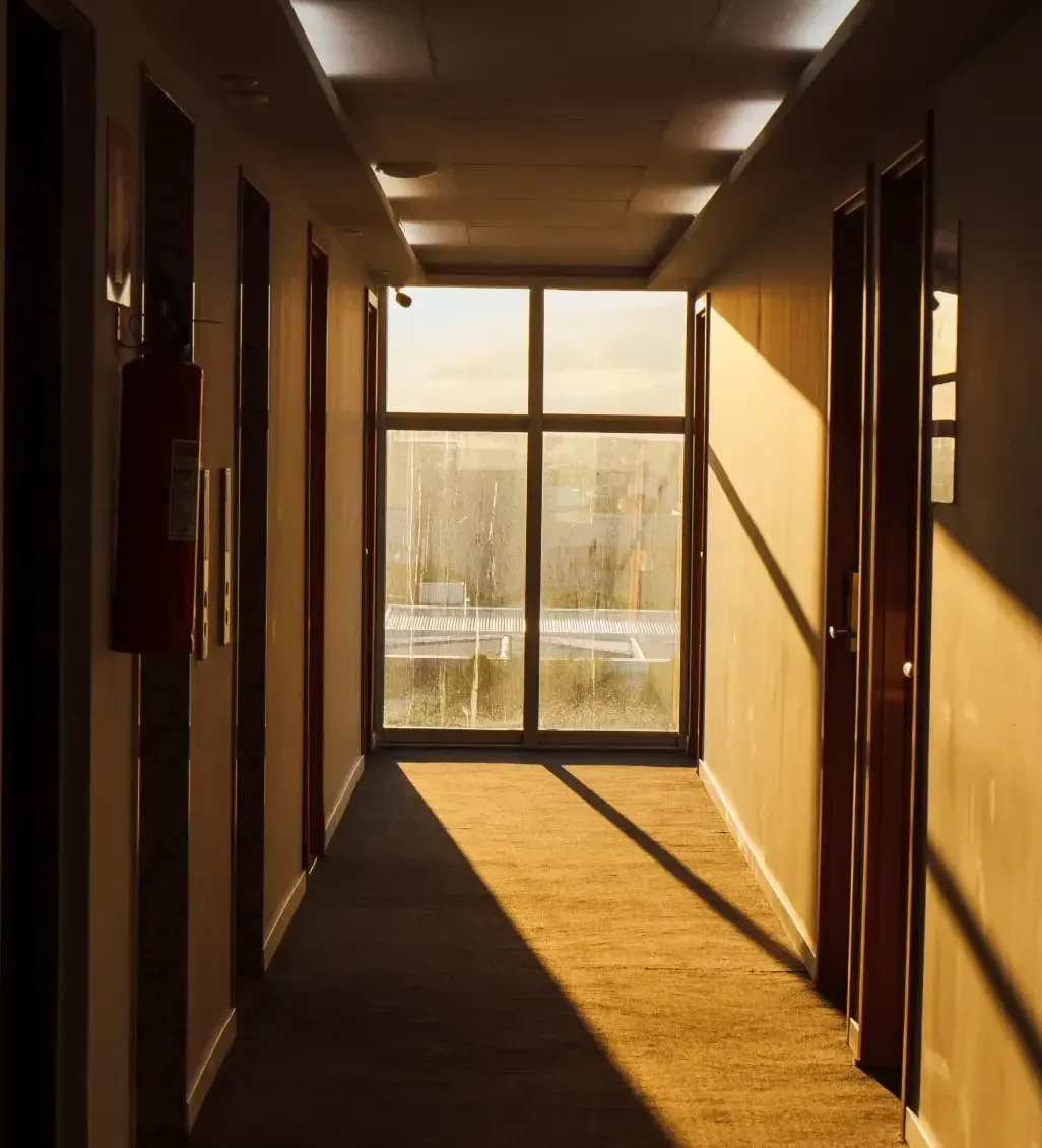 Corredor vazio e ensolarado com longas sombras, portas em ambos os lados e uma porta de vidro no final.