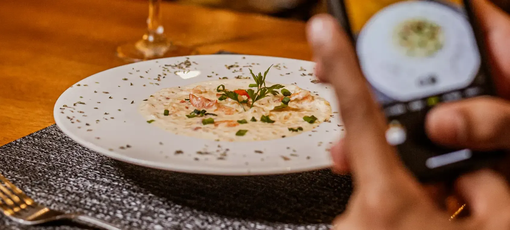 Mão segurando um celular, fotografando um prato em cima da mesa de um restaurante.