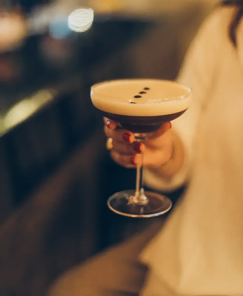 Mão segurando uma taça de coquetel com uma camada cremosa de espuma e grãos de café, em um bar com pouca luz.
