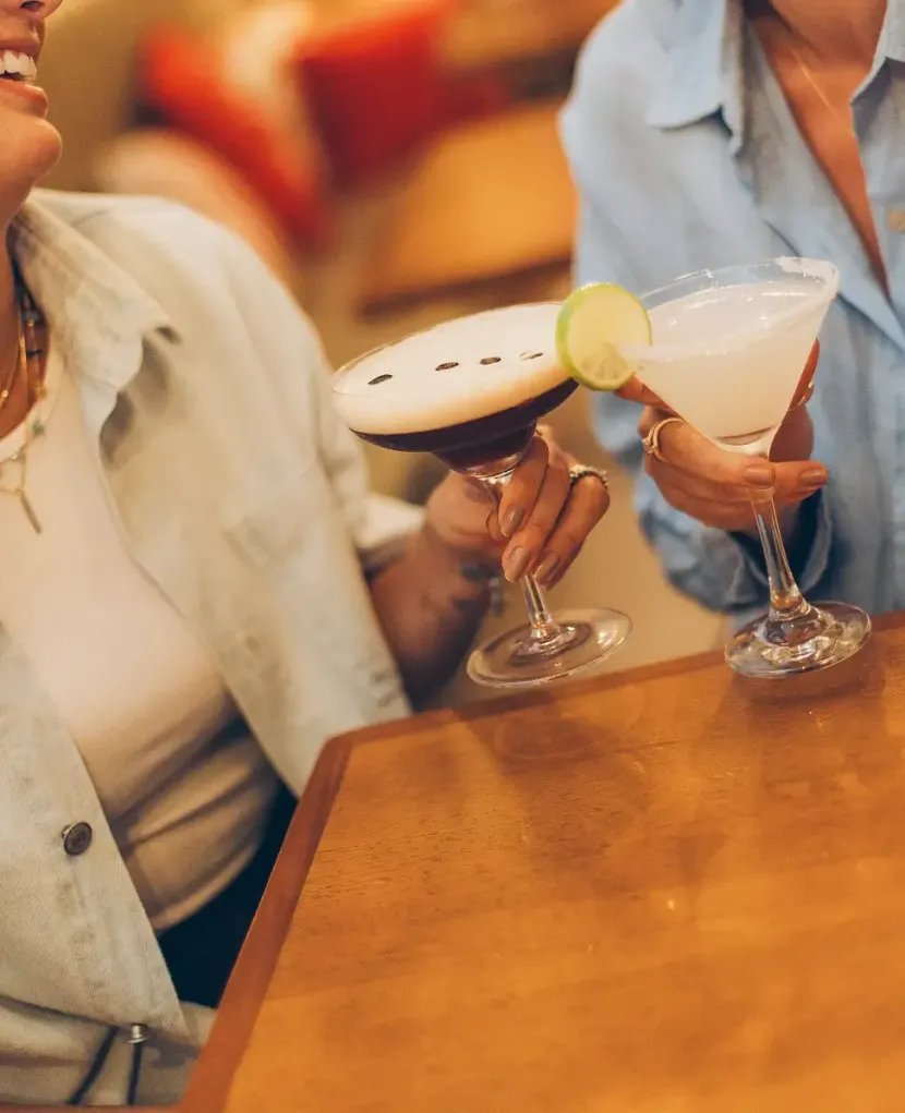 Duas pessoas segurando coquetéis com fatias de limão em uma mesa de madeira em um bar com iluminação aconchegante.